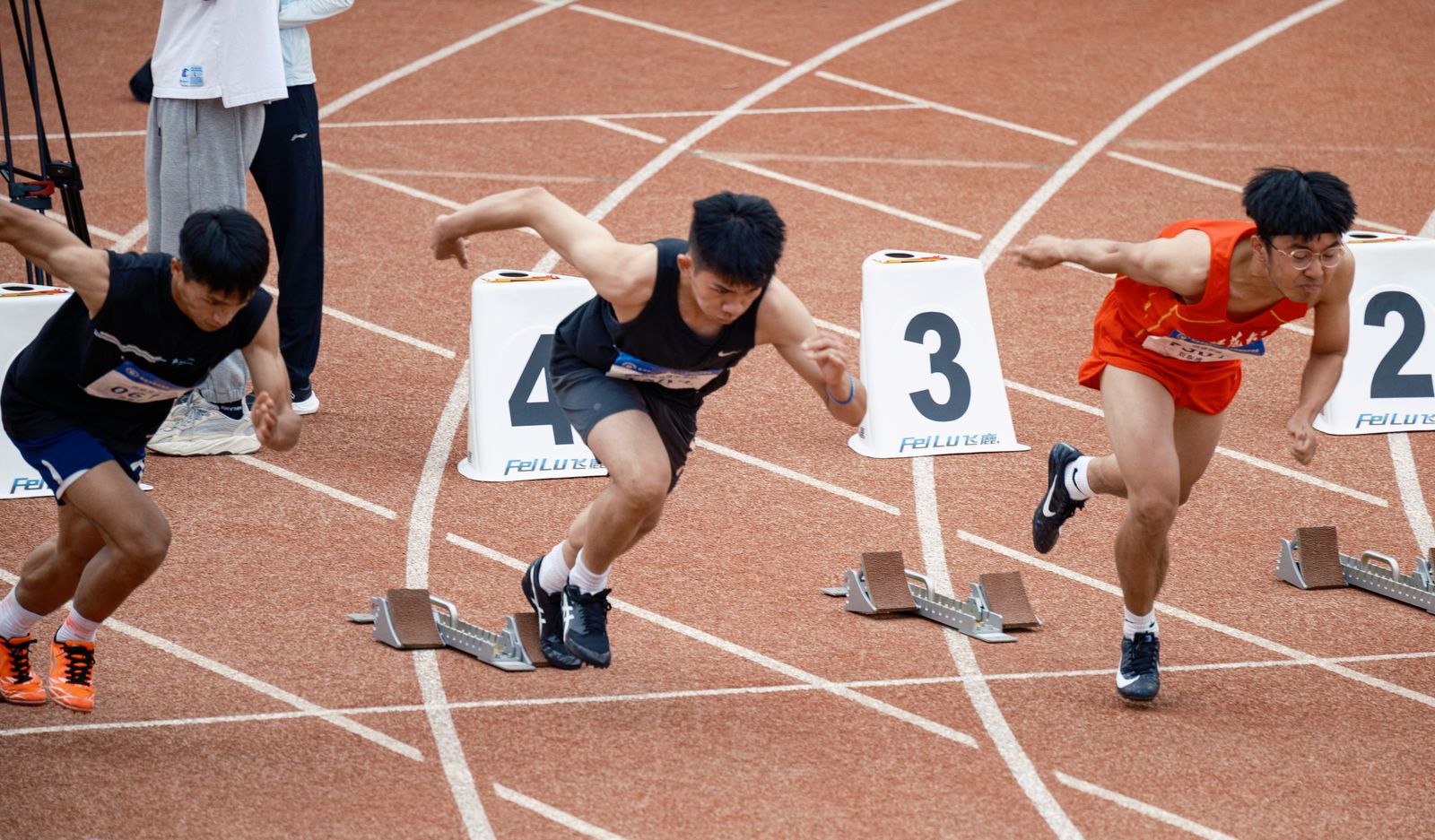 Corredores saindo dos blocos de largada numa pista de atletismo, um deles à frente